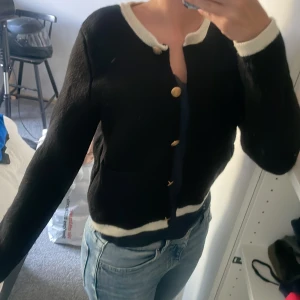 Svart kofta från Zara med guldknappar - Säljer en svart stickad kofta från Zara med kontrasterande vita kanter vid hals, ärmslut och nederkant. Koftan har snygga guldknappar framtill och två fickor. Perfekt till jeans för en chill men ändå uppklädd look. 
