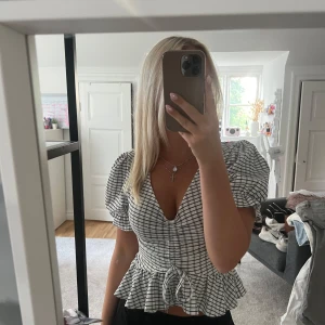 Vit och svart rutig blus med volang - Supersöt blus i vitt med svarta rutor, puffiga korta ärmar och v-ringning. Volang nertill och knyt i midjan ger en snygg siluett. Perfekt för dig som gillar trendiga detaljer och en feminin look.