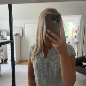 Vit blus med småprickigt mönster - Söt vit blus med små svarta prickar och korta ärmar. Blusen har v-ringning, knappar framtill och fina veckdetaljer vid bröstet. Lätt och luftig, perfekt för varmare dagar. Passar till både jeans och kjol för en fräsch look.