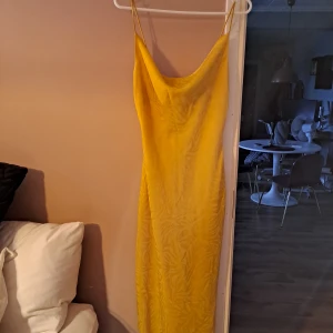 Gul midiklänning i sidenliknande material och tunna axelband - Snygg gul långklänning med tunna axelband och lätt draperad urringning. Klänningen har en slät, glansig yta och subtilt mönster i tyget som ger en extra touch. Perfekt för dig som vill sticka ut med färg och stil.