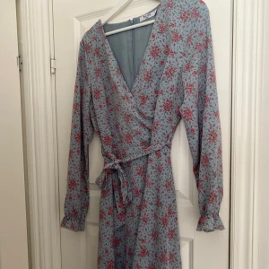 Blommig omlottklänning från NA-KD - Supersöt ljusblå omlottklänning från NA-KD med rosa blommigt mönster, långa ärmar med volangdetalj vid ärmslut och knytband i midjan. Perfekt för vår och sommar när du vill känna dig extra fin.