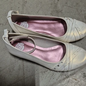 Ballerinaskor - Söta ballerinaskor från Melata med smal rem runt vristen och dekorativa detaljer framtill, inklusive små nitar och ett hjärta. Insidan är fodrad med glansigt rosa tyg som ger en extra touch. Perfekta för dig som gillar en feminin och stilren look.
