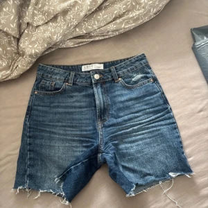 Blå jeansshorts från Primark - Snygga blå jeansshorts från Primark med hög midja och råa, fransiga benslut. Klassisk femficksmodell i denim med dragkedja och knapp framtill. Perfekta för sommaren och ger en avslappnad vibe.