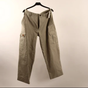 Beiga cargobyxor från Fjällräven - Snygga beiga cargobyxor från Fjällräven Ladies i 100% bomull. Byxorna har flera praktiska fickor med lock på sidorna och bak, samt klassisk cargomodell med raka ben. 46L