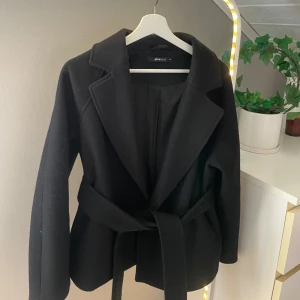 Svart kappa från Gina Tricot - Stilren svart kappa från Gina Tricot i storlek XS. Jackan har klassisk krage, lång ärm och knyts med bälte i midjan. 