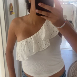Vit oneshoulder topp med volang Mango - Säljer en vit oneshoulder topp från Mango med bred volang och broderade hål längs kanten. Toppen är ärmlös och har en snygg asymmetrisk skärning. Perfekt till jeans eller kjol för en fräsch look.