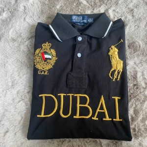 Svart pikétröja Polo Ralph Lauren Dubai - Svart pikétröja från Polo Ralph Lauren med stora broderier i gult och vitt, DUBAI-text framtill och nummer 2 på ryggen. Klassisk krage med vit rand, korta ärmar och detaljerad logga på bröstet. 100% bomull och custom fit. vid Snabb affär kan pris sänkas 