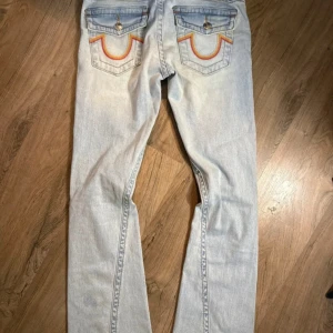 True Religion Joey lågmidjade bootcut jeans - Säljer ett par ljusblå True Religion Joey jeans med bootcut passform och låg midja. Jeansen har slitna detaljer vid knäna, klassiska bakfickor med färgglad brodyr och silverfärgade knappar. Bara använd några gånger så i mycket fint skick!💗