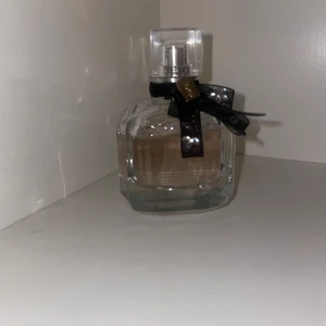 Burberry parfym med svart rosett - Elegant parfymflaska från Burberry i transparent glas med ljusrosa doftvätska. Flaskan har en fyrkantig form och pryds av en svart rosett med gulddetalj runt halsen. Perfekt för dig som gillar lyxiga och stilrena dofter.