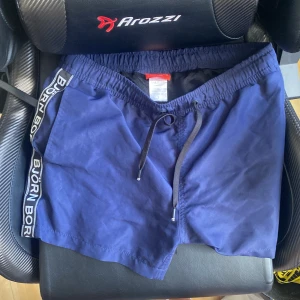 Blå Björn Borg badshorts med snörning - Snygga marinblå badshorts från Björn Borg med svart snörning i midjan och logga längs sidorna. Shortsen har elastisk midja och meshfoder för extra komfort. Perfekta för stranden eller poolen i sommar.