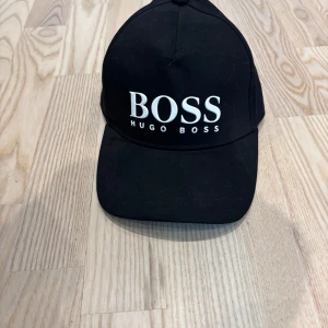 Svart keps från Hugo Boss - Svart keps från Hugo Boss med stor vit BOSS-logga framtill. Justerbar rem baktill med metallspänne där det också står BOSS. Kepsen är tillverkad i 100% bomull och har klassisk böjd skärm. Perfekt för en clean och stilren look.