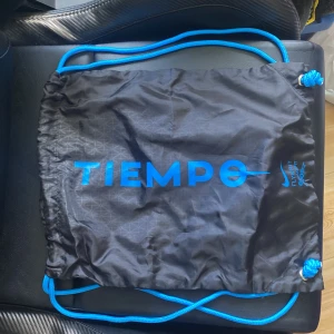 Nike Tiempo svart gympåse - Svart gympåse från Nike med blåa snören och blått tryck där det står TIEMPO. Väskan har subtilt geometriskt mönster och är perfekt för att bära träningskläder eller skor. Lätt och smidig att ta med till träningen.