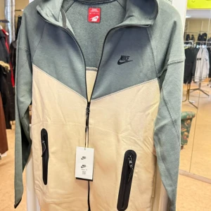 Nike beige och grå tech dress  - Finns alla storlekar!! Nike tech dress i snygg beige och grå färgblock-design. Jackan har hel dragkedja framtill, två svarta dragkedjefickor och Nike-logga på bröstet. Tillverkad i mjuk bomullsmix, perfekt för chill dagar eller när du är på språng. Långärmad och med sportig vibe.