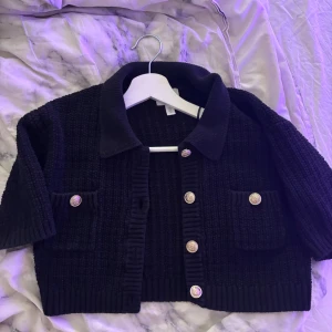 Svart stickad kofta med guldknappar - Snygg svart stickad kofta från H&M med croppad passform, stora guldiga knappar och två fickor framtill. Koftan har krage och ribbade detaljer vid ärmslut och nederkant. Perfekt för lager-på-lager och ger en chic vibe till din outfit.