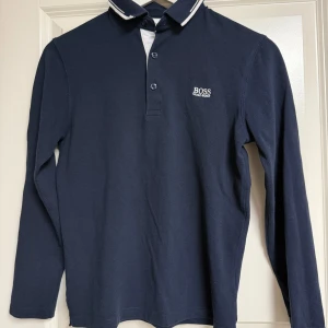 Marinblå långärmad piké från Hugo Boss - Snygg marinblå långärmad pikétröja från Hugo Boss med vit logga broderad på bröstet. Klassisk krage med vita detaljer och knappar framtill. Tillverkad i mjuk bomull som känns skön mot huden. Perfekt för dig som gillar stilrena och tidlösa plagg.