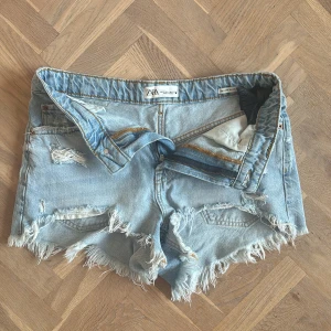 Ljusa jeansshorts från ZARA med slitningar - Säljer ett par ljusblå jeansshorts från ZARA med råa, fransiga kanter och flera slitningar för en riktigt edgy vibe. Klassisk femficksmodell med bälteshällor och dragkedja. Perfekta för sommardagar när du vill ha en avslappnad och cool stil.