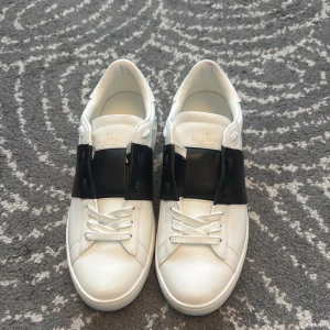 Valentino Garavani Open sneakers vit/svart - Snygga Valentino Garavani Open sneakers i vitt skinn med bred svart läderdetalj över vristen. Klassisk rund tå, vita snören och ikoniska nitar baktill på sulan. Perfekta för dig som gillar stilrena och lyxiga sneakers med edge.