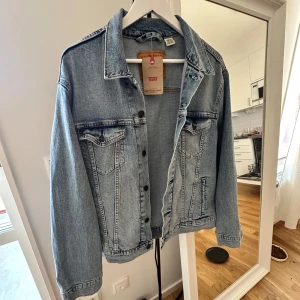 Levi's ljusblå jeansjacka L - Klassisk ljusblå jeansjacka från Levi's i modellen The Trucker Jacket. Jackan har standard fit, två bröstfickor med knappar och snygga sömdetaljer. Perfekt för dig som vill ha en tidlös och avslappnad look. Aldrig använd, lapp kvar.