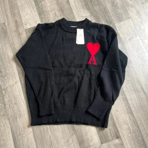 Svart stickad tröja från Ami Paris - Svart stickad tröja från Ami Paris med rund hals och ribbade muddar. På bröstet finns ett rött hjärta med bokstaven A, vilket ger en unik och trendig look. Tillverkad i bomull och passar perfekt till lager-på-lager-stil.