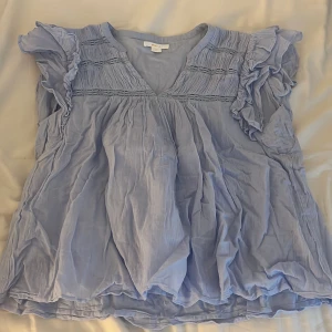 Ljusblå volangblus från Gina Tricot - Supersöt ljusblå blus från Gina Tricot med v-ringning, volangiga ärmar och fina broderade detaljer upptill. Luftig och flowy modell i tunt tyg, perfekt för varmare dagar. Ger en romantisk och chill vibe till din outfit. Säker i storlek 170 men passar även xxs/xs💞