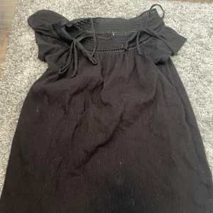 Svart offshoulder kortklänning - Svart kortklänning med offshoulder och smala axelband. Klänningen har snygga knytband och virkade detaljer upptill som ger en bohemisk känsla. Perfekt för dig som gillar en avslappnad men ändå trendig stil.