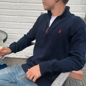 Mörkblå stickad tröja Polo Ralph Lauren - Mörkblå stickad tröja från Polo Ralph Lauren med halv dragkedja och ribbad krage. Klassisk röd logga broderad på bröstet. Långärmad modell med breda muddar och skön passform. Perfekt för kyliga sommarkvällar. 