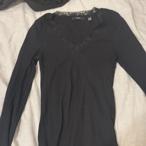 Svart ribbad topp med spets från Vero Moda - Svart långärmad topp från Vero Moda med ribbad struktur och v-ringning dekorerad med spetsdetaljer. Snygg och enkel att styla, perfekt för dig som gillar en stilren look med en touch av spets.