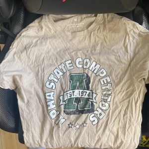 Beige t-shirt Alpha State Competitors - Beige t-shirt med tryck 'Alpha State Competitors' och stort grönt A på bröstet. Regular fit med rund hals och korta ärmar. Skön college-vibe och perfekt för dig som gillar sportig stil. Tillverkad i bomull.