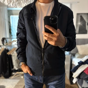 Mörkblå bomberjacka från Strömberg - Snygg mörkblå bomberjacka från Strömberg med dragkedja, ribbade muddar och coola detaljer på axlarna. Jackan har två fickor med dragkedja och en stilren krage med tryckknappar. Perfekt för dig som vill ha en clean och modern look.