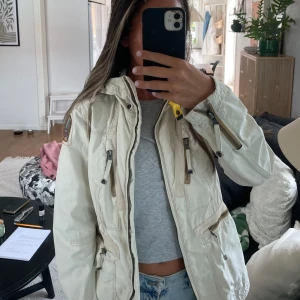 Parajumpers höstjacka - Snygg beige vindjacka från Parajumpers med luva, dragkedjor och små detaljer. Perfekt nu till hösten med en stilren och funktionell jacka till vardags. Storlek S