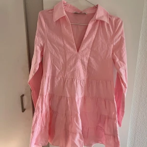 Rosa volangklänning med krage - Supersöt rosa kortklänning med volanger och lång ärm. Djup v-ringning och klassisk krage ger en chill men ändå trendig vibe. Perfekt för dig som gillar en luftig och flowy stil. Tillverkad i bomull för en skön känsla hela dagen.