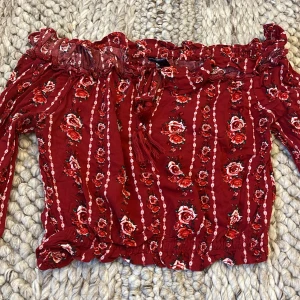 Röd blommig offshoulder topp H&M - Supersöt röd offshoulder topp från H&M med blommigt mönster och volangkant upptill. Toppen har långa ärmar, smockad resår vid ärmslut och midja samt knytband framtill. Den är croppad. Storlek 34