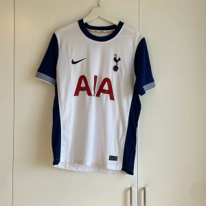 Tottenham Hotspur hemmatröja Nike S - Tottenham Hotspur hemmatröja från Nike i storlek S. Tröjan är vit med mörkblå detaljer på ärmar och krage, samt röd AIA-logga på bröstet. Tillverkad i lätt och ventilerande polyester, perfekt för match eller träning. Klassisk klubbmärke på bröstet.