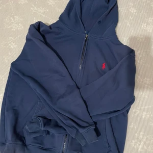 Marinblå hoodie från Polo Ralph Lauren - Snygg marinblå hoodie från Polo Ralph Lauren med dragkedja och huva. Tröjan har det klassiska röda broderade logotypen på bröstet och är i bomull. Perfekt för en chill och stilren look.