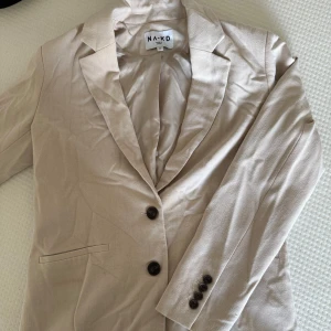 Beige kavaj från NA-KD, storlek 34 - Stilren beige kavaj från NA-KD i storlek 34. Klassisk modell med två knappar framtill, fickdetaljer och snyggt skräddad passform.