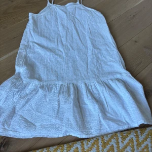 Vit somrig klänning med volang - Superfin vit klänning med tunna axelband och volang nertill. Lätt strukturerat tyg som känns luftigt och perfekt för varma dagar. Enkel och stilren modell som ger en avslappnad vibe.