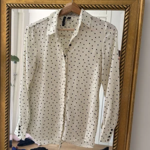 Vit skjorta med svarta hjärtan från Mango - Supersöt vit skjorta från Mango med små svarta hjärtan över hela tyget. Klassisk krage, knäppning framtill och långa ärmar med knappar vid ärmslut. Perfekt för dig som gillar lekfulla detaljer men ändå vill ha en stilren look.