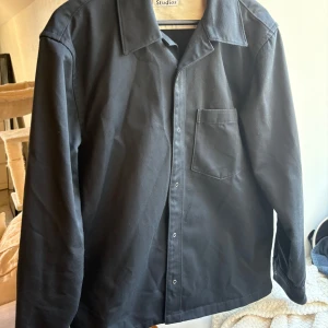 Svart overshirt från Acne Studios - Stilren svart overshirt från Acne Studios i en mix av bomull och polyester. Skjortan har klassisk krage, tryckknappar framtill och en bröstficka. Perfekt för lager-på-lager och enkel att matcha med det mesta. Väldigt bra skick