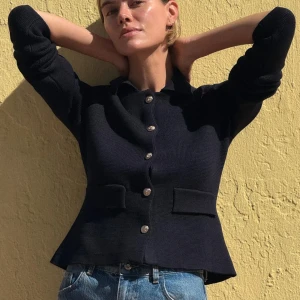 Svart stickad cardigan jacka från Zara - Svart stickad cardigan från Zara med snygga guldknappar framtill och två fickor. Jackan har en klassisk krage och figurnära passform som ger en clean och stilren look. Perfekt att slänga över en t-shirt eller klänning.