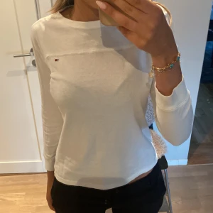 Vit långärmad tröja Tommy Hilfiger - Vit långärmad tröja från Tommy Hilfiger med liten broderad logga på bröstet. Enkel och stilren design med rund halsringning. Perfekt till jeans eller kjol för en clean look. Barn storlek 12-14 år. 