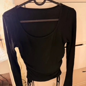 Svart långärmad topp med snörning - Svart långärmad topp med rund halsringning och snygga snörningsdetaljer i sidorna som ger en trendig look. Tunt, ribbat material som sitter tajt och framhäver figuren. Perfekt att styla med jeans eller kjol för en cool vardagsoutfit.