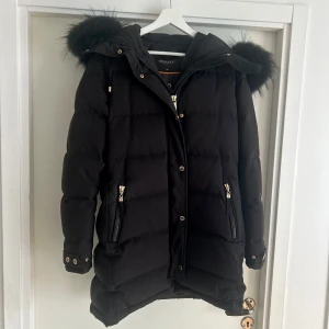 Svart dunjacka med päls från Hollies - Snygg svart dunjacka från Hollies med fluffig päls på huvan. Jackan har guldiga dragkedjor, knappar och två fickor framtill. Quiltad design och längre baktill för extra stilpoäng. Perfekt för kalla vinterdagar och riktigt varm. Har använts enstaka gånger, som ny.                                  Jackan passar även storlek 40&38