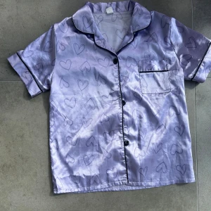Lila satinpyjamas-skjorta med hjärtan - Superfin lila satinpyjamas-skjorta från Shein med svarta hjärtmönster och svarta kantdetaljer. Skjortan har korta ärmar, bröstficka och knäppning framtill. Perfekt för dig som gillar mjuka och glansiga material med gulliga detaljer. Nypris var 30kr och aldrig använd eller sovit i. Pris kan diskuteras 