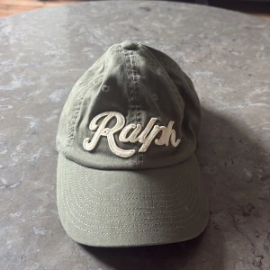 Olivgrön keps från Polo Ralph Lauren - Snygg olivgrön keps från Polo Ralph Lauren med broderad 'Ralph' framtill och 'Polo' baktill. Justerbar rem i bak för perfekt passform. Kepsen är i bomull och har en klassisk, avslappnad look som passar till det mesta.