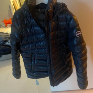 Mörkblå dunjacka från Colmar med huva, strl 140 - Snygg svart vinterdunjacka från Colmar med glansig finish och vadderad design. Jackan har huva, dragkedja framtill och Colmar-logga på ärmen. Perfekt för kalla dagar och ger en cool, sportig vibe. (Litet hål på en armen så därmed har jag sänkt priset)