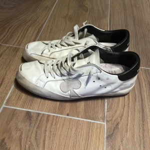 Vita sneakers The Gallerist med klöver - Snygga vita sneakers från The Gallerist med svart hälparti och coolt klövermotiv på sidan. Skorna har snörning, platt sula och rund tå. Tillverkade i skinn med svarta detaljer och text på hälen. Perfekta för dig som gillar stilrena men unika sneakers. Pris kan diskuteras!