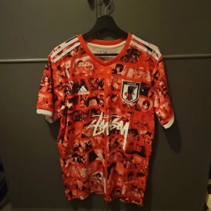 Japan Adidas x Stussy anime fotbollströja - Unik Japan fotbollströja från Adidas i samarbete med Stussy, täckt av röda och vita anime-motiv. Tröjan har korta ärmar, V-ringning och klassiska Adidas-ränder på axlarna. Materialet är lätt och andas, perfekt för dig som gillar streetwear och anime. 