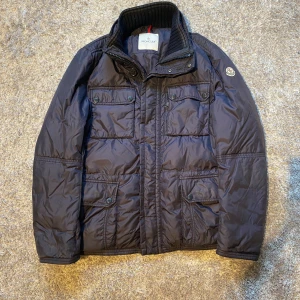 Svart Moncler pufferjacka herr - Snygg svart pufferjacka från Moncler med hög krage och flera fickor framtill. Jackan har quiltad design, dragkedja och knappar samt Moncler-logga på ärmen. Perfekt för kalla dagar och ger en clean streetstyle-vibe.