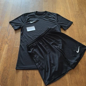 Svart Nike fotbollströja och shorts - Säljer ett svart fotbollskit från Nike med Dri-FIT-teknologi. Tröjan är kortärmad och har en vit Nike-logga på bröstet. Matchande svarta shorts med vit Nike-logga. Perfekt för träning eller match.