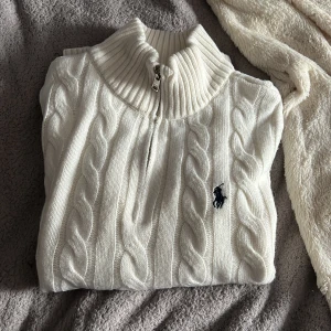 Vit stickad tröja från Ralph Lauren - Säljer en stilren vit stickad tröja från Ralph Lauren med kabelstickat mönster och dragkedja vid halsen. Tröjan har långa ärmar och en broderad logotyp på bröstet. Perfekt för kyliga dagar!